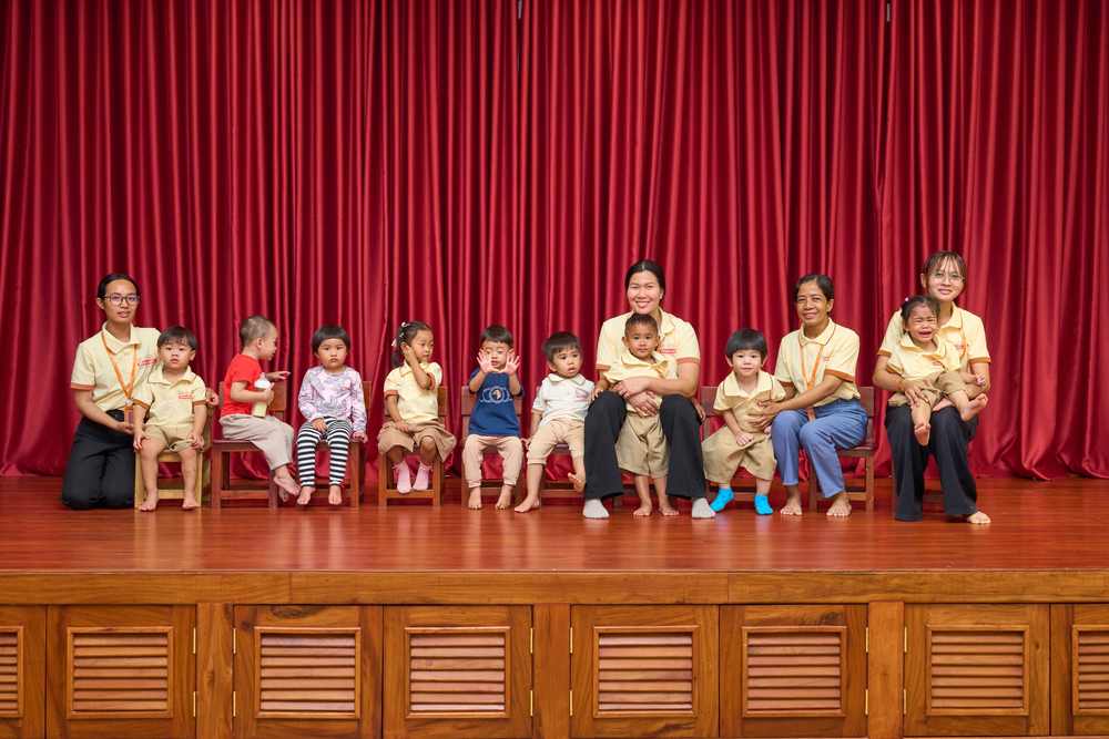 Bunny Class seated on small chairs on the hall stage with their teachers beside them, red curtain behind