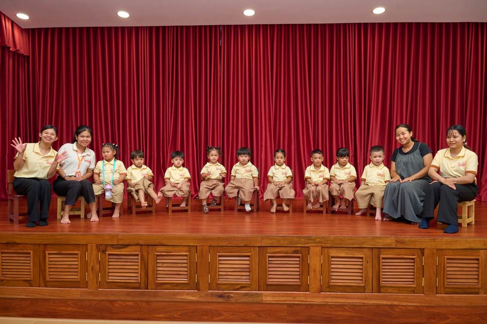 Panda Class in a row of small chairs on the hall stage, a teacher seated at each end
