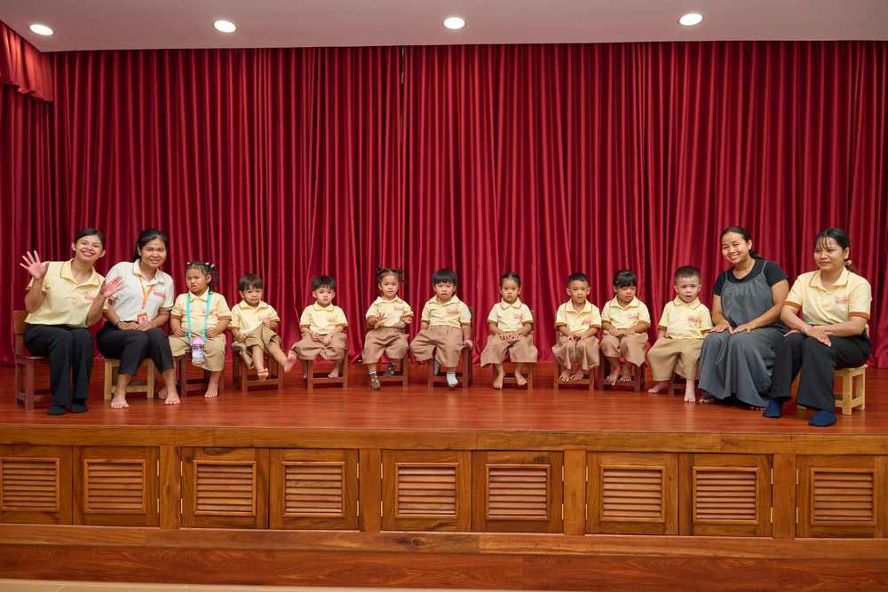 Panda Class in a row of small chairs on the hall stage, a teacher seated at each end