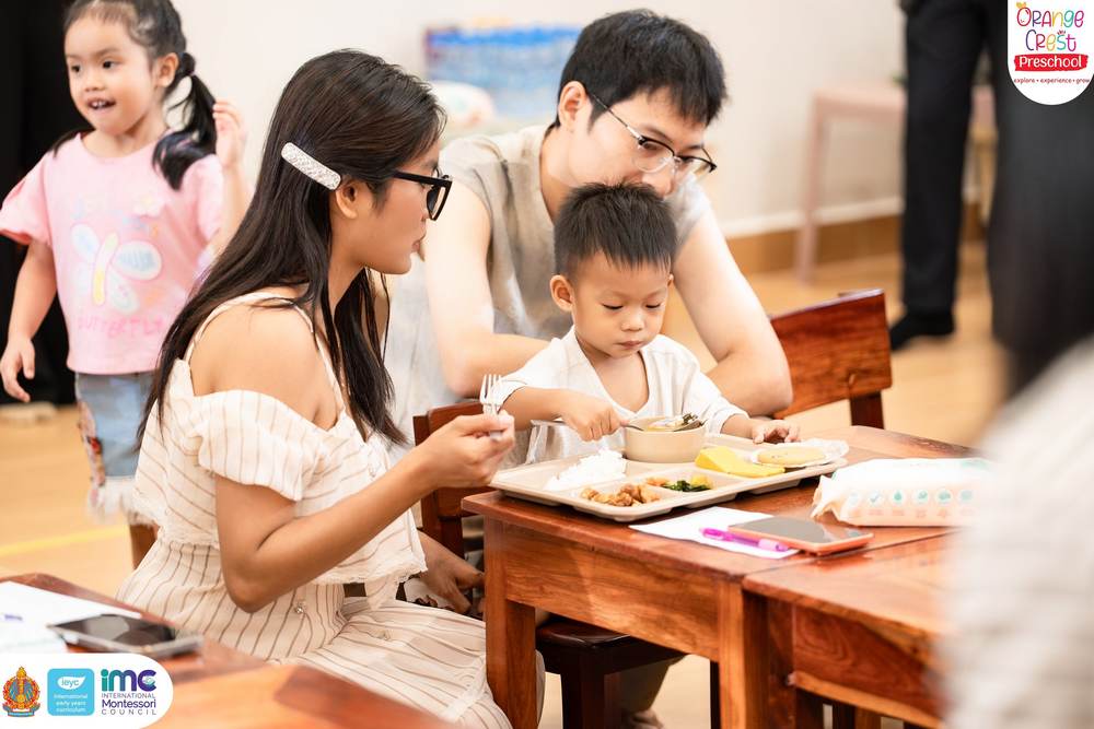 A mother and father sitting either side of their small son at a wooden table, helping him eat from a school lunch tray