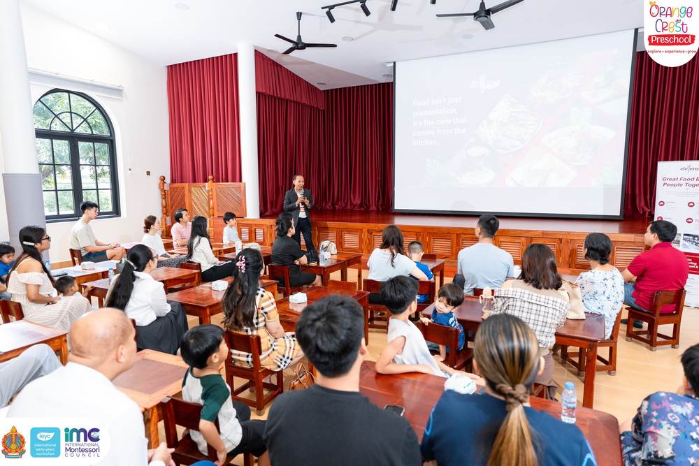 Parents and children seated at the long wooden tables in the school hall, facing a presentation screen