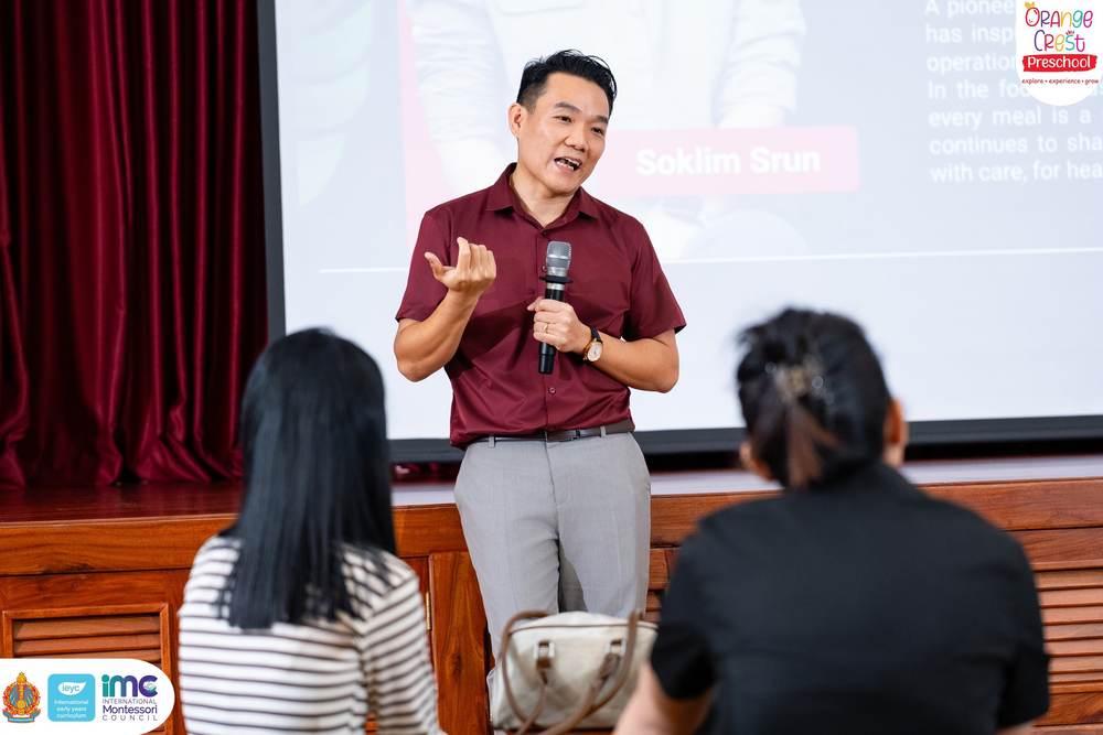A representative of Eleven One Kitchen speaking with a microphone in front of the presentation screen in the school hall