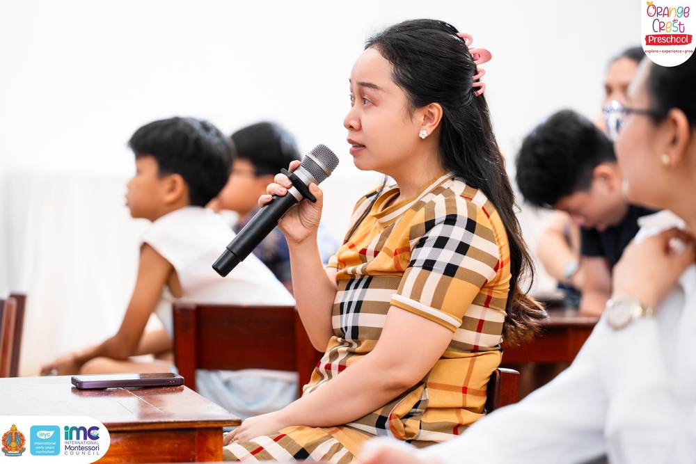 A mother speaking into a handheld microphone from her seat in the hall, her child sitting beside her