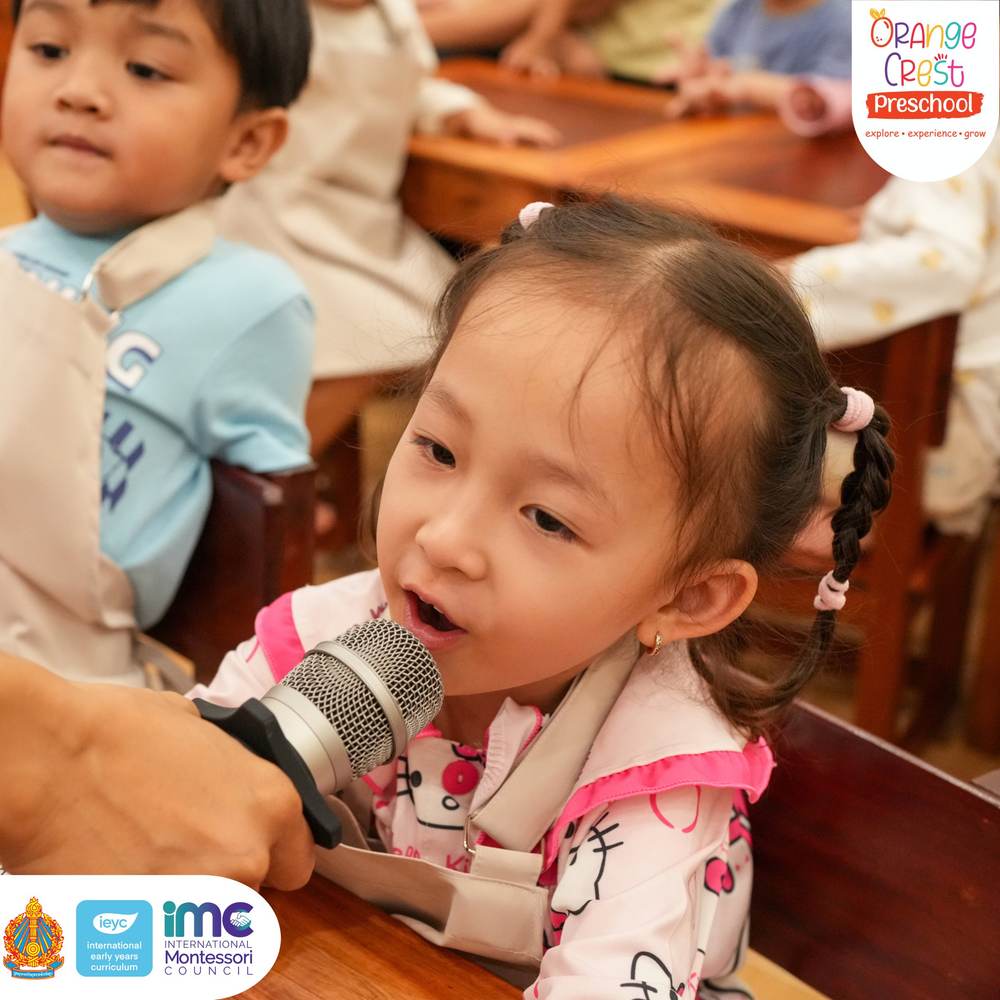 A small girl speaking into a handheld microphone held in front of her, classmates watching
