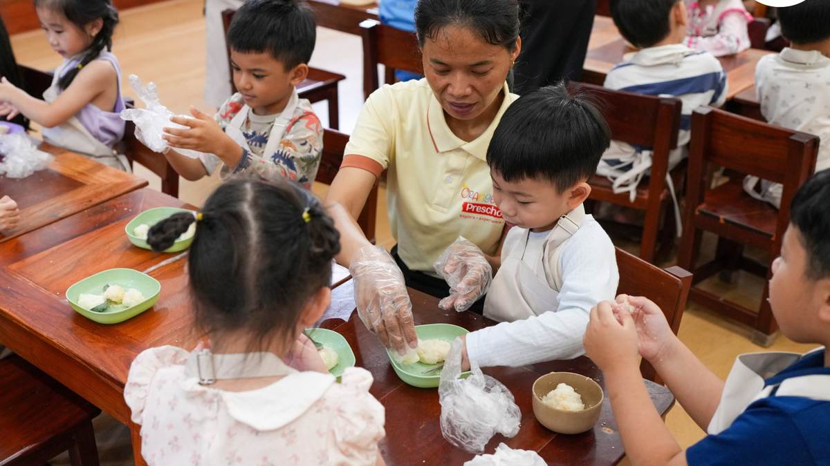 A teacher at Orange Crest Preschool helping children shape Bay Ben rice balls at a long wooden table, their hands in gloves and bowls of rice in front of them