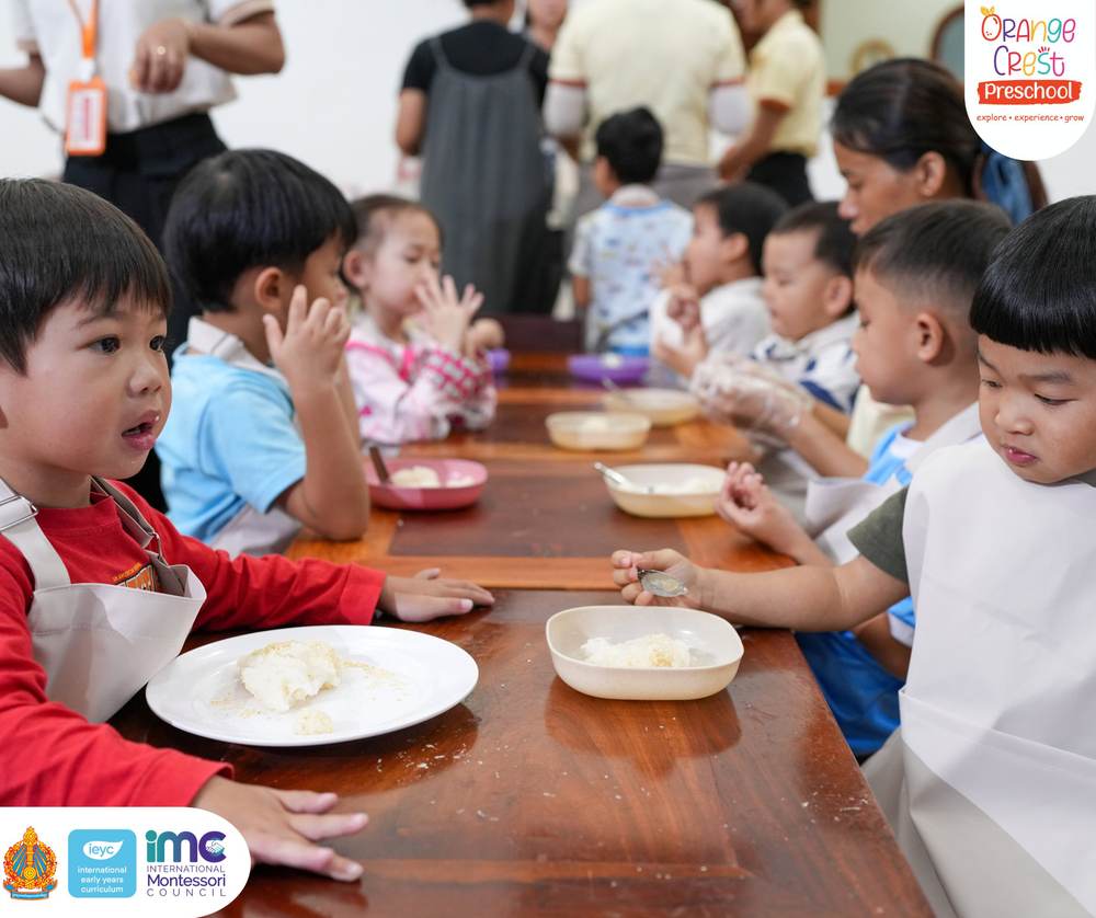 The long wooden table lined with children in aprons, each with a plate and a bowl of rice