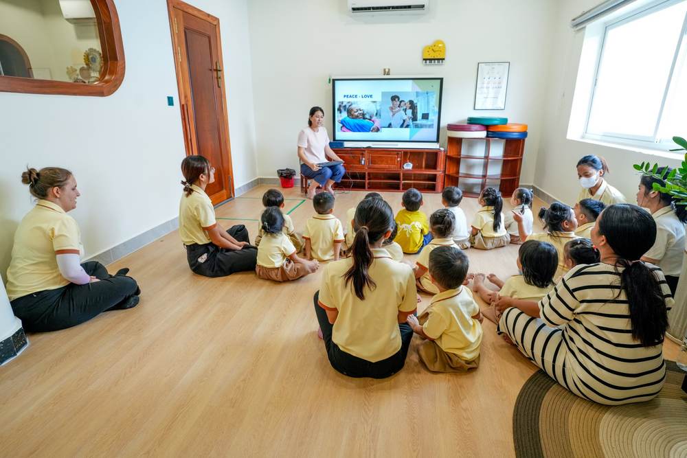 Kindergarten children and their teachers sitting together on the classroom floor, facing a screen that shows the words Peace – Love and a picture of a family