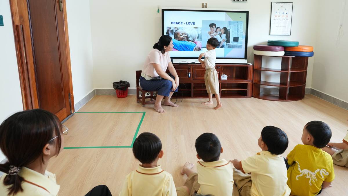 Peace Day at Orange Crest kindergarten in Phnom Penh: a child in a yellow uniform stands at the classroom screen showing the words Peace – Love, a teacher seated alongside, while classmates watch from the floor