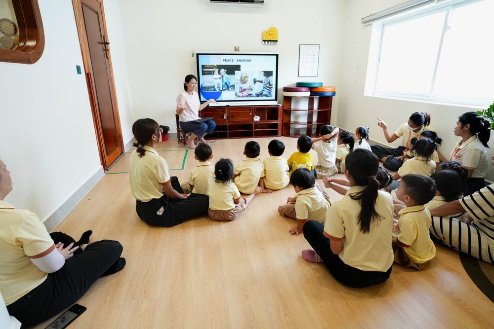 A teacher presents the slide Peace – Sharing to kindergarten children sitting on the floor, with more teachers sitting among them