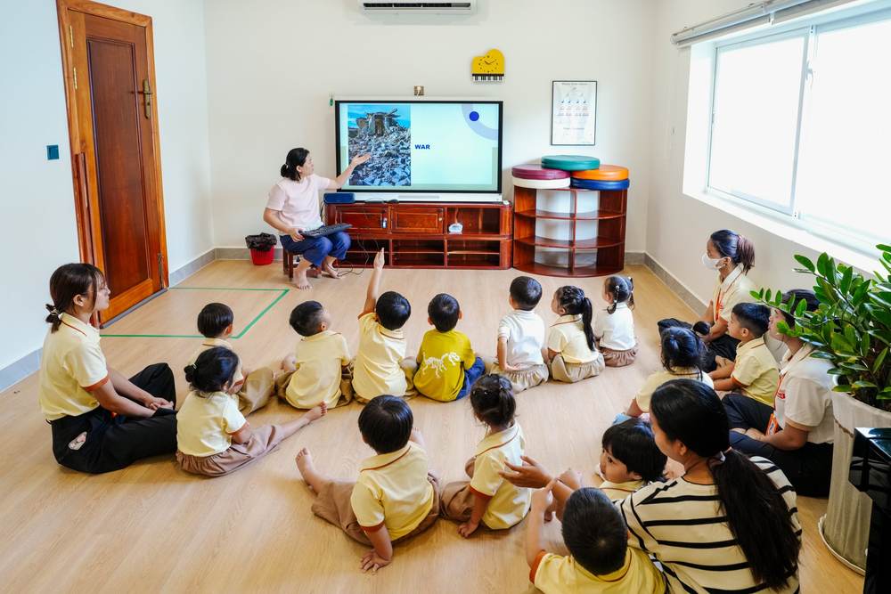 A kindergarten teacher points to a picture of war on the classroom screen while children sitting on the floor raise their hands to answer