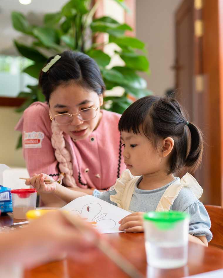 A teacher sitting beside a child at a low table, guiding them through an art activity