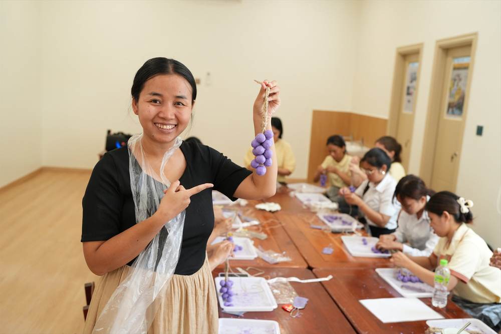 A teacher holding up her finished bunch of purple grapes and smiling, with colleagues still working at the table behind her
