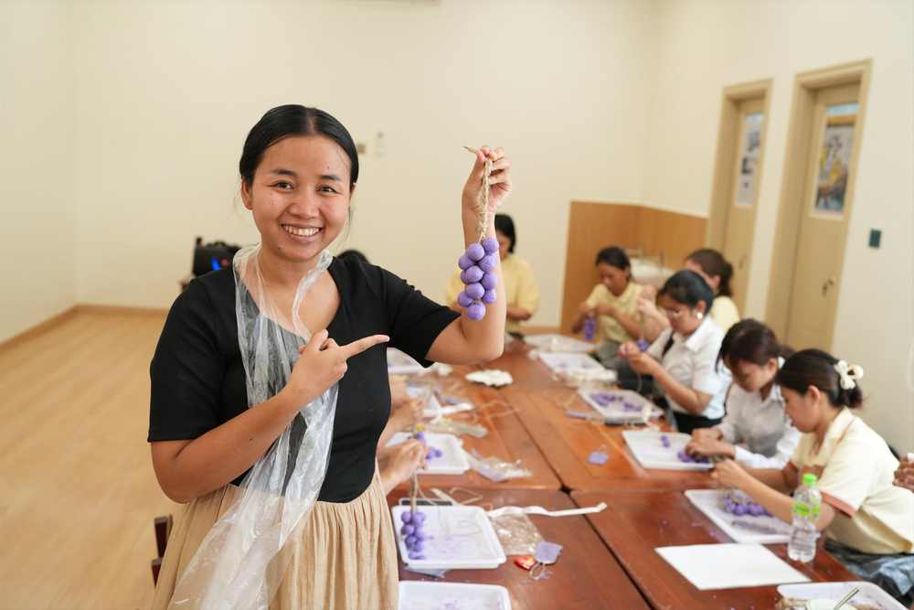 A teacher holding up her finished bunch of purple grapes and smiling, with colleagues still working at the table behind her