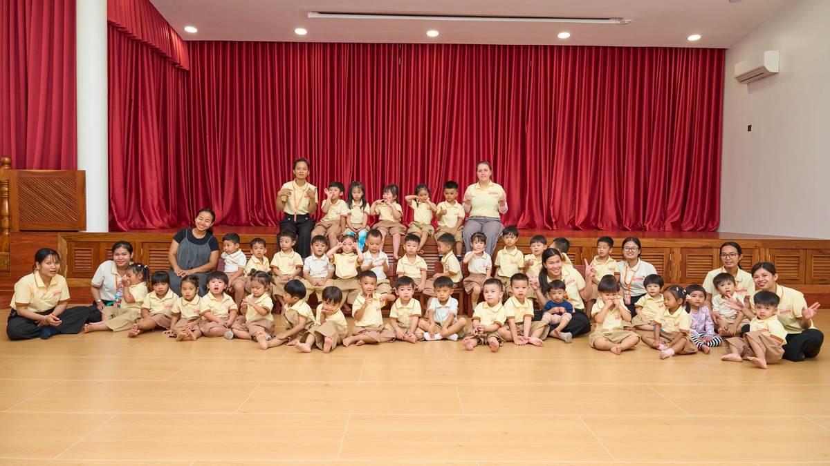 The whole of Orange Crest Preschool, children in yellow polo shirts and their teachers, seated together on the hall floor in front of the stage and its red curtain