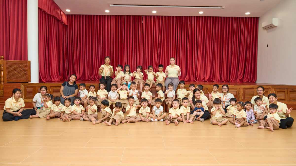 The whole of Orange Crest Preschool, children in yellow polo shirts and their teachers, seated together on the hall floor in front of the stage and its red curtain